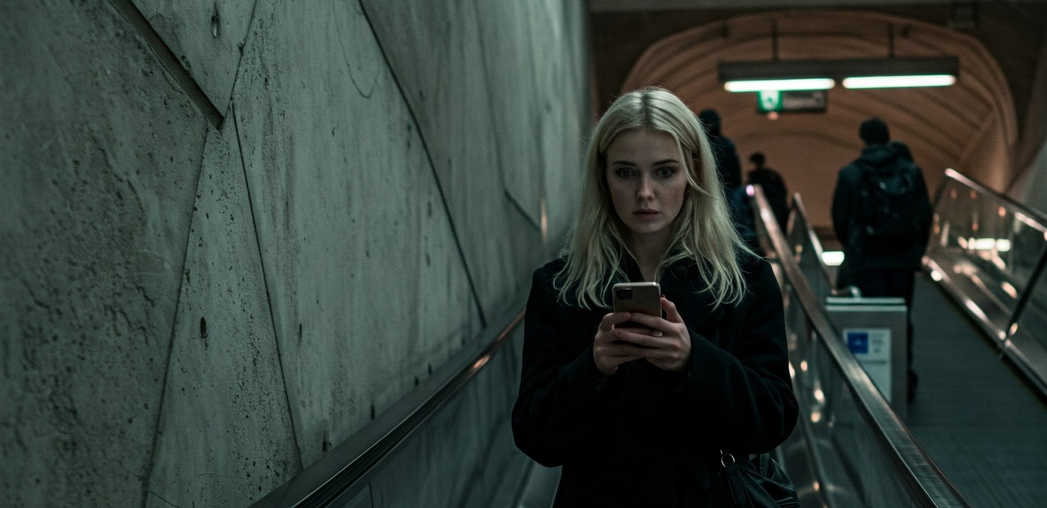 a blonde woman in a black coat riding up an escalator in a modern, urban metro station. She is looking at her smartphone.